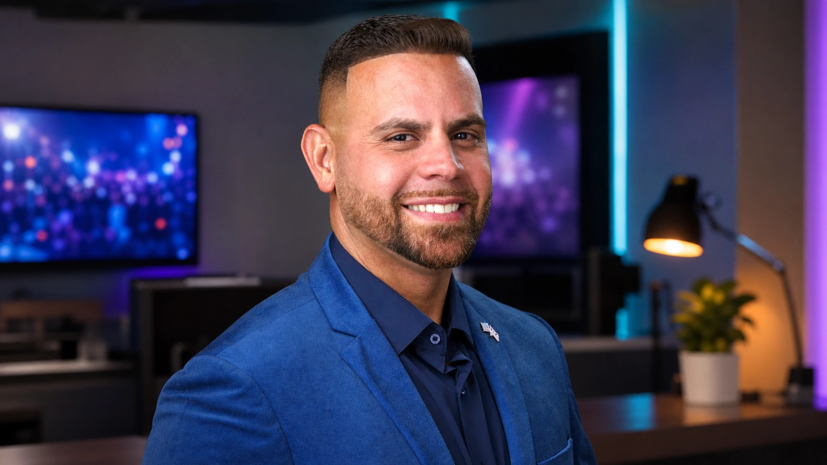 Carlos Vazquez smiling toward the camera in his blue blazer and flag lapel pin in an event broadcast office
